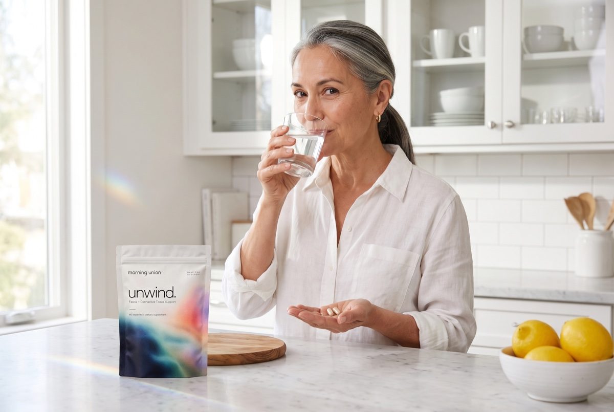 A woman in her late 50s taking two capsules with water in her kitchen, unwind. on the counter